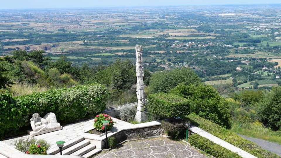 Terrazza panoramica su una collina con pavimentazione decorata, gradini e aiuole fiorite, affacciata su una vasta pianura con campi e aree boschive. Al centro si vede un alto monumento in pietra e una scultura bianca vicino al parapetto.