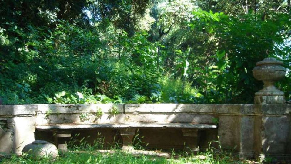 Area verde ombreggiata con fitta vegetazione e alberi sullo sfondo, davanti a un basso muretto in pietra con una lunga panca integrata. Ai lati del muretto sono presenti elementi decorativi in pietra, tra cui un vaso/urna e un piedistallo con coppa.