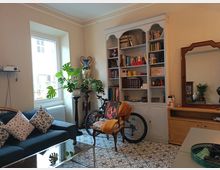 Soggiorno luminoso con grande finestra, pavimento a piastrelle decorate e divano scuro con cuscini. Sulla parete c’è una libreria bianca piena di libri e oggetti, con una bicicletta appoggiata davanti e, a lato, un comò in legno con grande specchio.