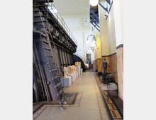 Corridoio interno di un edificio tecnico o museo industriale, con una grande macchina metallica con scaletta e passerella lungo la parete sinistra e modelli esposti su piedistalli. A destra si vedono pareti rivestite in marmo, lampade a globo e una macchina d’epoca, con pavimento a mosaico e una scala sul fondo.