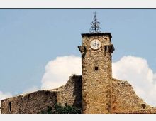 Torre campanaria in pietra con orologio circolare sulla facciata e croce in ferro battuto in cima, integrata in un complesso di mura e tetti in pietra. Sullo sfondo si vede il cielo azzurro con nuvole e vegetazione alla base della struttura.