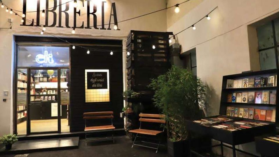 Ingresso di una libreria in un cortile interno, con grande insegna “LIBRERIA” e porta a vetri illuminata che mostra scaffali di libri all’interno. Nell’area esterna ci sono fili di luci sospese, panchine in legno, una pianta in vaso e un espositore con libri su un tavolo.