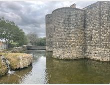 Mura di una fortificazione in pietra scura con torri cilindriche che si affacciano su un fossato d’acqua. A sinistra si vede una sponda erbosa con un piccolo salto d’acqua, alberi e una passerella in legno sotto un cielo nuvoloso.