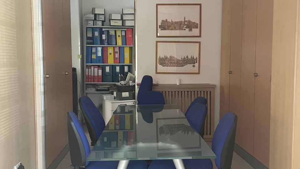 Piccola sala riunioni in ufficio con tavolo rettangolare in vetro e sedie girevoli blu, circondata da armadi in legno. In fondo si vede un’area con scaffali pieni di raccoglitori colorati, due stampe incorniciate sulla parete e faretti incassati nel soffitto.