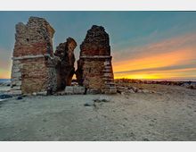 Rovine di una struttura in mattoni e pietra, con due pilastri parzialmente crollati, su una spiaggia sabbiosa vicino al mare. Sullo sfondo si vede la linea dell’orizzonte e un cielo al tramonto con strisce di colore arancione e blu.