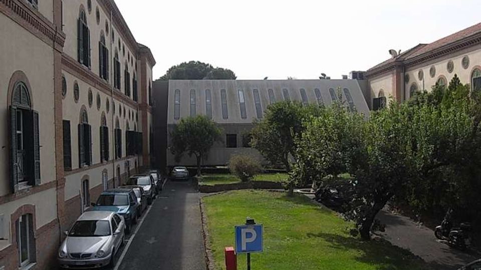 Cortile interno tra edifici in stile storico con facciate chiare e finestre ad arco con persiane, affacciato su una strada stretta con auto parcheggiate. Al centro c’è un’area verde con alberi e un cartello di parcheggio, chiusa sul fondo da un edificio moderno con finestre verticali.