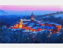 Veduta panoramica di un borgo collinare al crepuscolo, con tetti coperti di neve e un campanile illuminato al centro del paese. Sullo sfondo si vedono colline e montagne in lontananza sotto un cielo rosa e azzurro.