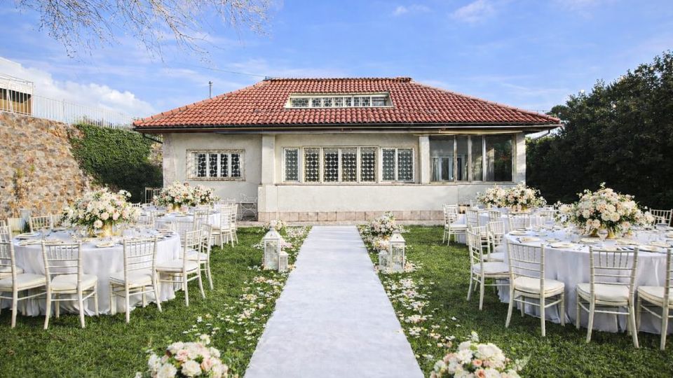 Giardino allestito per un ricevimento con tavoli rotondi coperti da tovaglie bianche e sedie chiare, disposti ai lati di una passerella centrale decorata con petali e lanterne. Sullo sfondo si vede una villa a un piano con tetto in tegole rosse e grandi finestre, circondata da mura in pietra e vegetazione.
