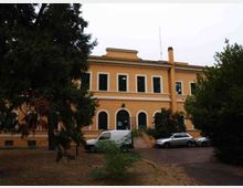 Edificio a due piani color ocra con finestre rettangolari al piano superiore e ingressi ad arco al piano terra, con una breve scalinata centrale. Davanti si vede un vialetto con auto parcheggiate e un’area con alberi e vegetazione che incorniciano la facciata.