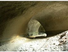 Interno di una grotta o tunnel scavato nella roccia, con volta arrotondata e pareti grezze. In fondo si vede un piccolo passaggio ad arco illuminato, con pavimento terroso e detriti.
