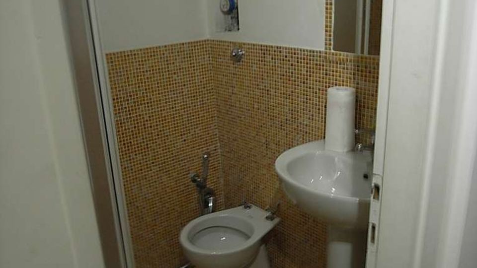 Piccolo bagno con pareti e pavimento in mosaico giallo, con wc e lavabo a colonna accanto a uno specchio. A sinistra si vede una cabina doccia semicircolare con ante in vetro.