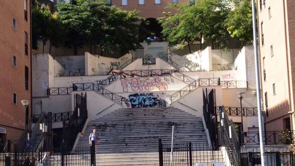 Ampia scalinata urbana terrazzata con rampe laterali e ringhiere metalliche, incassata tra alti edifici in mattoni. Muri bianchi con graffiti e alberi in cima incorniciano i diversi livelli collegati dalle scale.
