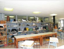 Interno di un laboratorio artistico con lunghi tavoli da lavoro in legno e sgabelli, sotto un soffitto con luci fluorescenti. Lungo le pareti ci sono scaffalature metalliche piene di sculture e modelli in vari materiali, con finestre e pareti vetrate sullo sfondo.