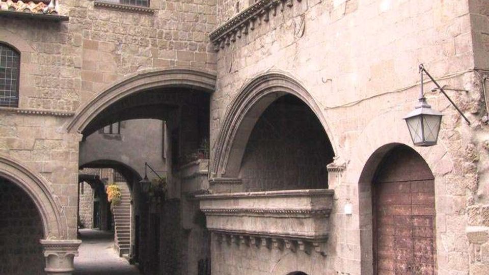 Cortile di un edificio storico in pietra, con portici ad archi e passaggi coperti che collegano le facciate. Si vedono finestre ad arco con inferriate, un grande portone in legno con scalini e una lanterna a muro su una parete in muratura.