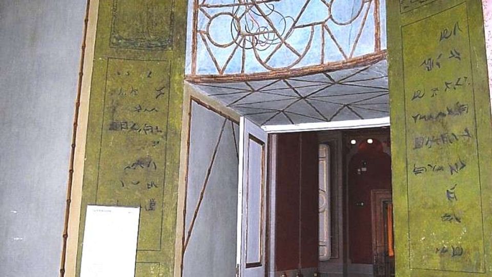 Interno di un edificio storico con grande portale decorato, pareti verdi con pannelli geometrici e fregi dipinti. Sopra l’ingresso un affresco con un uccello entro motivi ornamentali, che conduce a una sala adiacente con pavimento scuro e sedie lungo la parete.