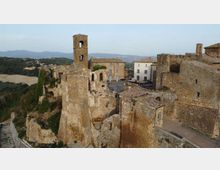 Veduta panoramica di un borgo medievale in pietra con resti di mura e una torre campanaria, affacciato su uno sperone roccioso. Sullo sfondo si vedono colline e campagna, con case compatte attorno a una piccola piazza.