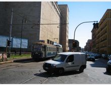 Strada urbana con binari del tram al centro, attraversata da una rete di cavi aerei e semafori. Lungo la via si vedono un grande edificio moderno in pietra e palazzi a più piani, con traffico di veicoli e un tram in corsa.