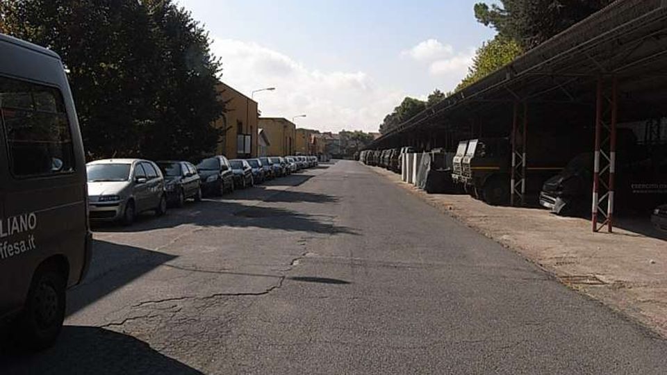 Strada asfaltata ampia in un’area industriale, con auto parcheggiate in fila sul lato sinistro e una lunga tettoia metallica con veicoli e materiali stoccati sul lato destro. Alberi e lampioni costeggiano la strada, che si estende in prospettiva verso edifici bassi sullo sfondo.