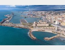 Veduta aerea di una città costiera con porto e moli, con navi ormeggiate e infrastrutture industriali sullo sfondo. In primo piano si vede un promontorio con un grande complesso fortificato in pietra e strade costiere che delimitano baie e frangiflutti sul mare.