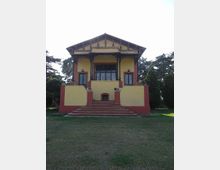 Edificio a due piani in stile villa, con facciata gialla e dettagli rosso mattone, portico centrale con colonne decorative e grande scalinata frontale. La struttura è circondata da un prato e da alberi alti sullo sfondo.