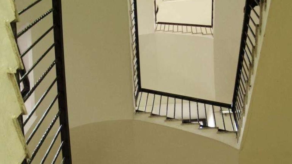 Vano scala interno di un edificio, ripreso dal basso verso l’alto, con rampe sovrapposte e ringhiere metalliche scure che incorniciano un’apertura centrale rettangolare. Pareti chiare e soffitto ad arco, con finestra in fondo che illumina lo spazio.