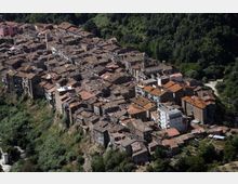 Vista aerea di un borgo collinare con case in pietra addossate tra loro e tetti in coppi, disposto lungo un crinale. Il centro abitato è circondato da fitta vegetazione e pendii boscati.