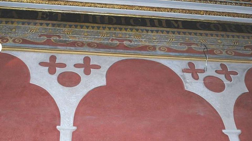 Dettaglio di una parete interna decorata con archi stilizzati rossi e colonne bianche, sormontata da fasce ornamentali policrome a motivi geometrici e iscrizioni. La parte superiore mostra cornici e fregi con mosaico o pittura decorativa.