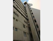 Veduta dal basso di un alto edificio multipiano in cemento, con facciate grigie e bianche e numerose finestre. Al centro si nota un grande vano verticale tipo torre tecnica o ascensore esterno che sale fino al tetto, con cielo visibile in alto tra le pareti.