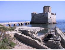 Fortezza in pietra sul mare con torre quadrata e mura merlate, collegata alla riva da un ponte in pietra ad archi. In primo piano si vedono rovine di murature e scogli lungo una piccola insenatura con acqua calma.