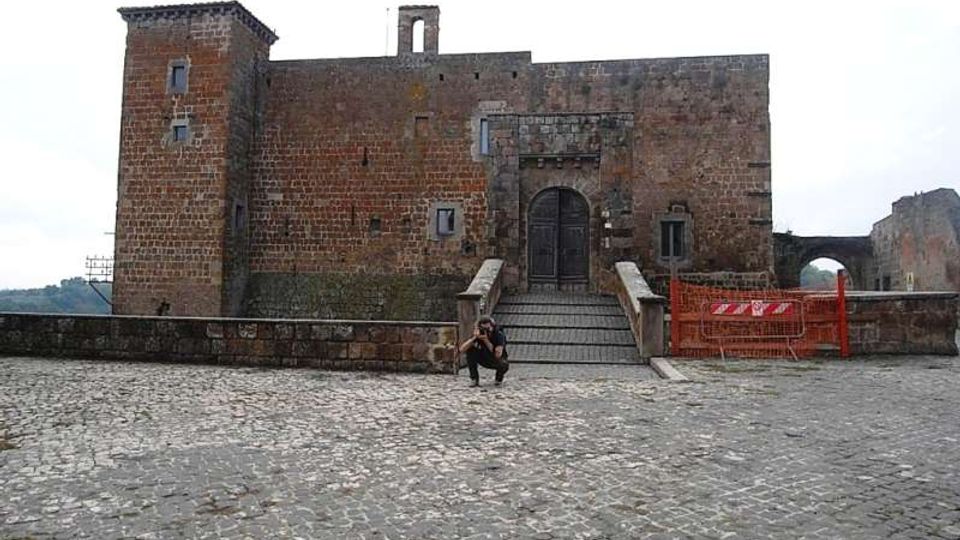 Facciata di un castello in pietra e mattoni con torre laterale e grande portale ad arco, raggiungibile tramite una scalinata con parapetti. In primo piano si vede un ampio piazzale lastricato in pietra, con una zona transennata sul lato destro.