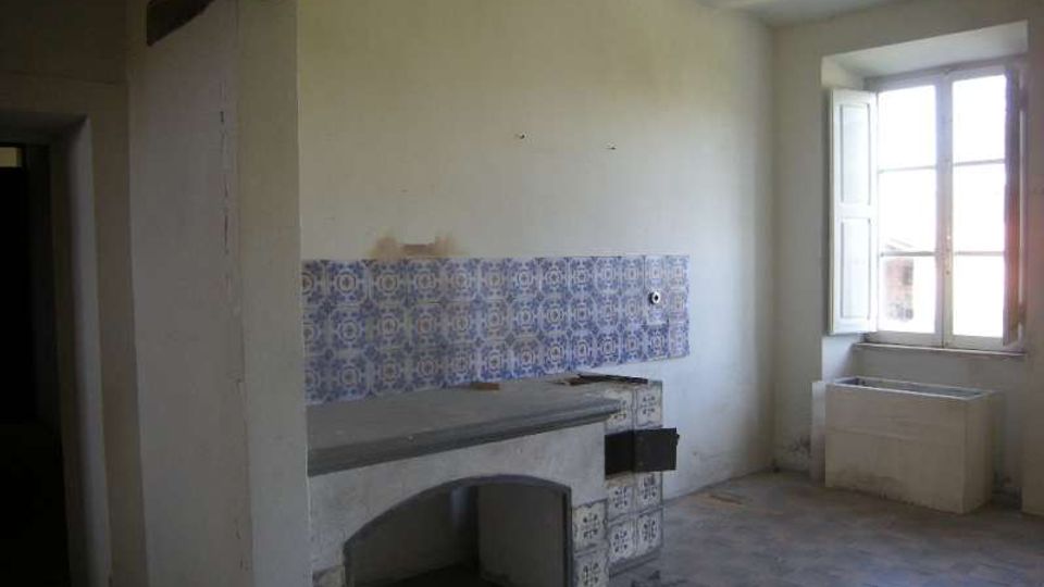 Interno di una cucina rustica e spoglia con pareti bianche, pavimento in piastrelle e una mensola in muratura rivestita di piastrelle decorative blu. A destra c’è una grande finestra con scuri aperti e, sotto, un lavello in muratura.