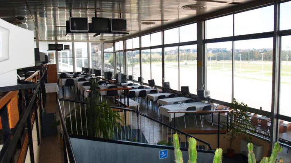 Interno di una sala ristorante o lounge con grandi vetrate a tutta altezza lungo un lato, tavoli apparecchiati e sedie disposte in file. Soffitto metallico con schermi sospesi, passerella con ringhiera e piante in vaso, con vista esterna su un’ampia area aperta.