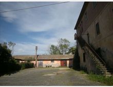Cortile esterno in cemento davanti a edifici rurali, con un basso fabbricato intonacato e tetto a falda sullo sfondo e un edificio più alto a destra con scala metallica esterna. Sullo sfondo si vedono alberi e un cielo parzialmente nuvoloso, con cavi elettrici sospesi.
