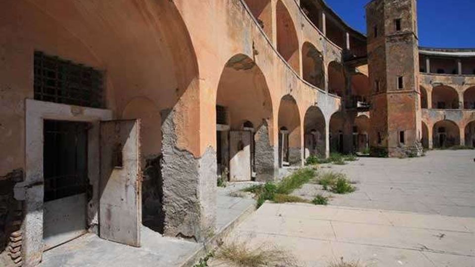 Cortile interno di un complesso storico in rovina, con porticati ad archi su più livelli e intonaco scrostato. Ampio pavimento in cemento con ciuffi d’erba, porte e aperture murate lungo le arcate e una torre in muratura sullo sfondo sotto cielo sereno.