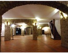 Ampio interno in stile rustico con pavimento in cotto e colonne in pietra che sorreggono una serie di archi, illuminati da appliques a parete. Sulla destra si vede una scala in legno che sale al piano superiore e, sullo sfondo, un’area con tavoli e sedie sotto le volte.