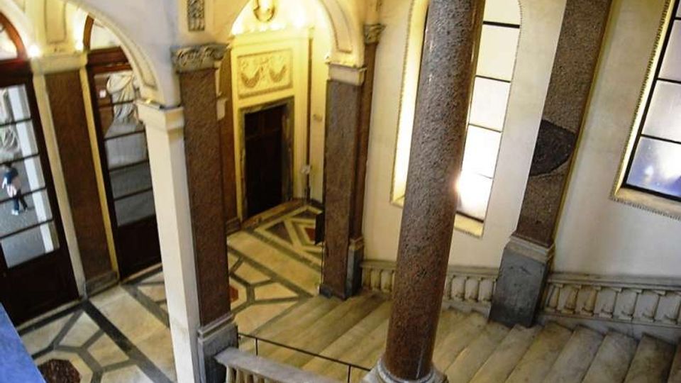 Interno di un atrio storico con scale in pietra e pavimento a motivi geometrici in marmo, illuminato da grandi finestre. Colonne massicce e archi decorati incorniciano porte in legno e un ingresso con cornice ornamentale.