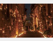 Vicolo stretto in pietra tra edifici in mattoni e intonaco, illuminato di notte da numerose candele in piccoli lumini appesi alle pareti e disposti lungo il percorso. La strada in lieve discesa è fiancheggiata da finestre, porte e vasi con piante, creando un corridoio luminoso tra le facciate.