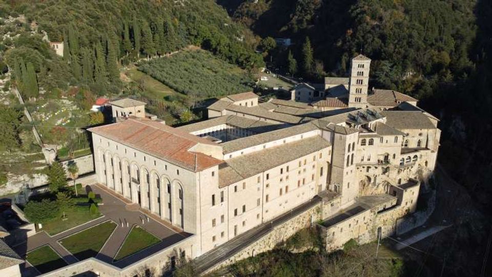 Veduta aerea di un grande complesso monastico in pietra con tetti in coppi, chiostro interno e una torre campanaria, costruito su un pendio con mura di contenimento. Intorno si vedono boschi e campi coltivati, con viali e piccoli edifici annessi.