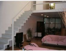 Interno di un loft moderno con scala bianca a vista che conduce a un soppalco con ringhiera metallica. Soggiorno open space con divano basso, TV, pavimento in legno e parete vetrata sul fondo che separa un’area con piante e arredi.