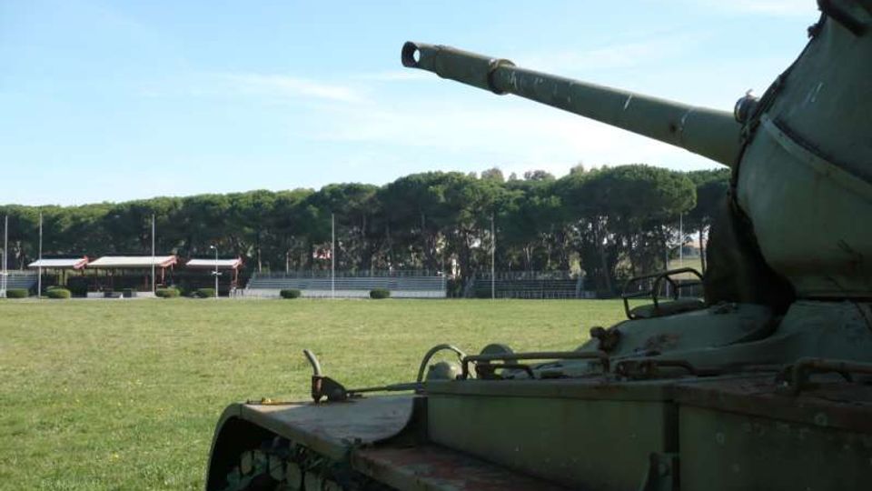 Campo erboso all’aperto con alberi sullo sfondo e una tribuna o edificio basso in lontananza. In primo piano si vede parzialmente un carro armato con cannone rivolto verso sinistra.