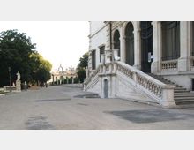Ampio viale lastricato accanto a un edificio storico in pietra chiara, con grandi arcate e cancellate; una scalinata monumentale con balaustra conduce all’ingresso sopraelevato. A sinistra si vede un’area alberata con statue lungo il percorso e, sullo sfondo, strutture architettoniche simili a un portico.