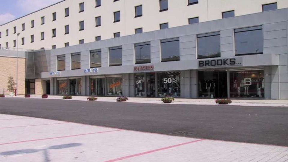 Edificio commerciale moderno a più piani con facciata chiara e file regolari di finestre, con un porticato al piano terra e vetrine di negozi con insegne. Davanti si vede un’ampia area pavimentata e asfaltata con alcune fioriere lungo il marciapiede.