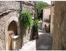 Vicolo stretto in un borgo storico, con pavimentazione in pietra e alti muri di edifici in muratura. A sinistra si vede un portale ad arco in pietra e piante in vaso lungo il muro, mentre il passaggio scende verso case con balconi e vegetazione sullo sfondo.