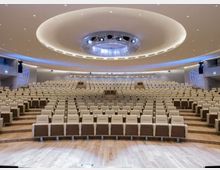 Ampio auditorium moderno con file di poltrone beige disposte a gradoni e pavimento in legno. Il soffitto curvo presenta un grande elemento circolare con illuminazione integrata e pannelli laterali con proiezioni luminose.