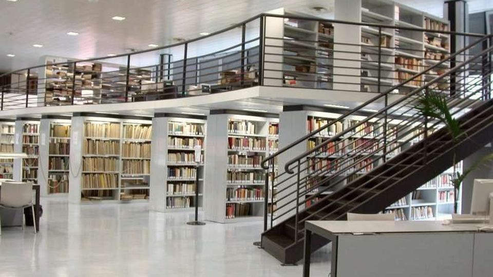 Interno di una biblioteca moderna a due livelli, con ampie scaffalature di libri e un soppalco con ringhiere metalliche. Una scala in metallo collega i piani in un ambiente luminoso con pavimento chiaro e arredi essenziali.