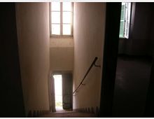 Vano scala interno stretto con pareti chiare, gradini in discesa e corrimano metallico a destra. Una porta aperta al piano inferiore e una finestra alta illuminano lo spazio, con un’altra finestra laterale dotata di persiane verdi.