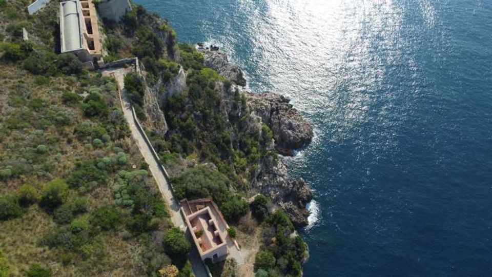 Veduta aerea di un promontorio roccioso sul mare con vegetazione mediterranea, una strada stretta e alcuni edifici in muratura lungo la costa. Il litorale presenta scogliere a picco e acque blu con riflessi di luce.