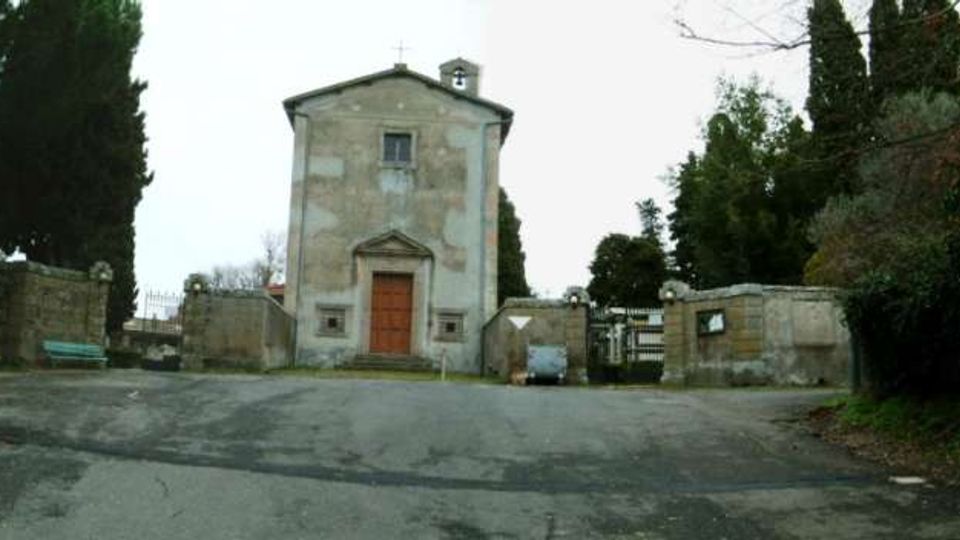 Piccola cappella in muratura intonacata con portale centrale e campanile a vela, situata all’ingresso di un cimitero con muri in pietra e cancelli laterali. Davanti si apre uno spiazzo asfaltato, circondato da cipressi e vegetazione.