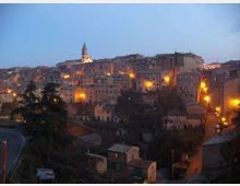 Panorama al crepuscolo di un borgo collinare con case in pietra addossate tra loro e un campanile che spicca sul profilo degli edifici. Luci stradali arancioni illuminano le facciate e le vie, con una strada curva e alberi in primo piano.