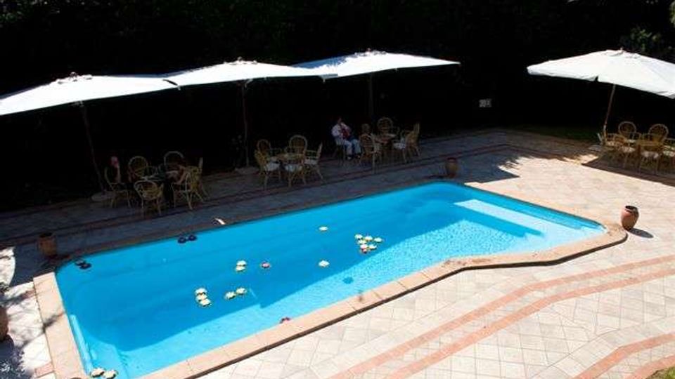 Piscina rettangolare all’aperto con bordo in pietra e pavimentazione in piastrelle geometriche, circondata da un’area solarium. Lungo un lato ci sono grandi ombrelloni bianchi con tavolini e sedie, con una siepe alta sullo sfondo.